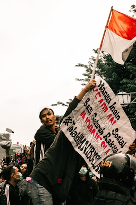 A group of people gathered in a protest. One individual is prominently holding a sign with Indonesian text and a red-and-white Indonesian flag. The crowd appears to be animated and engaged, with some wearing face masks and others making gestures. A police helmet is visible in the crowd.