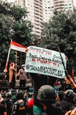 A group of people engaged in a street protest, with one person holding a large banner with text. The crowd includes individuals wearing helmets and uniforms. In the background, there are tall buildings and trees, and a person holds an Indonesian flag.