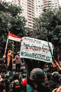 A group of people engaged in a street protest, with one person holding a large banner with text. The crowd includes individuals wearing helmets and uniforms. In the background, there are tall buildings and trees, and a person holds an Indonesian flag.