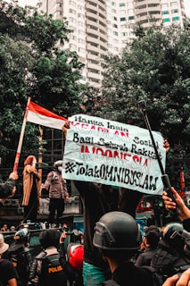 A group of people engaged in a street protest, with one person holding a large banner with text. The crowd includes individuals wearing helmets and uniforms. In the background, there are tall buildings and trees, and a person holds an Indonesian flag.