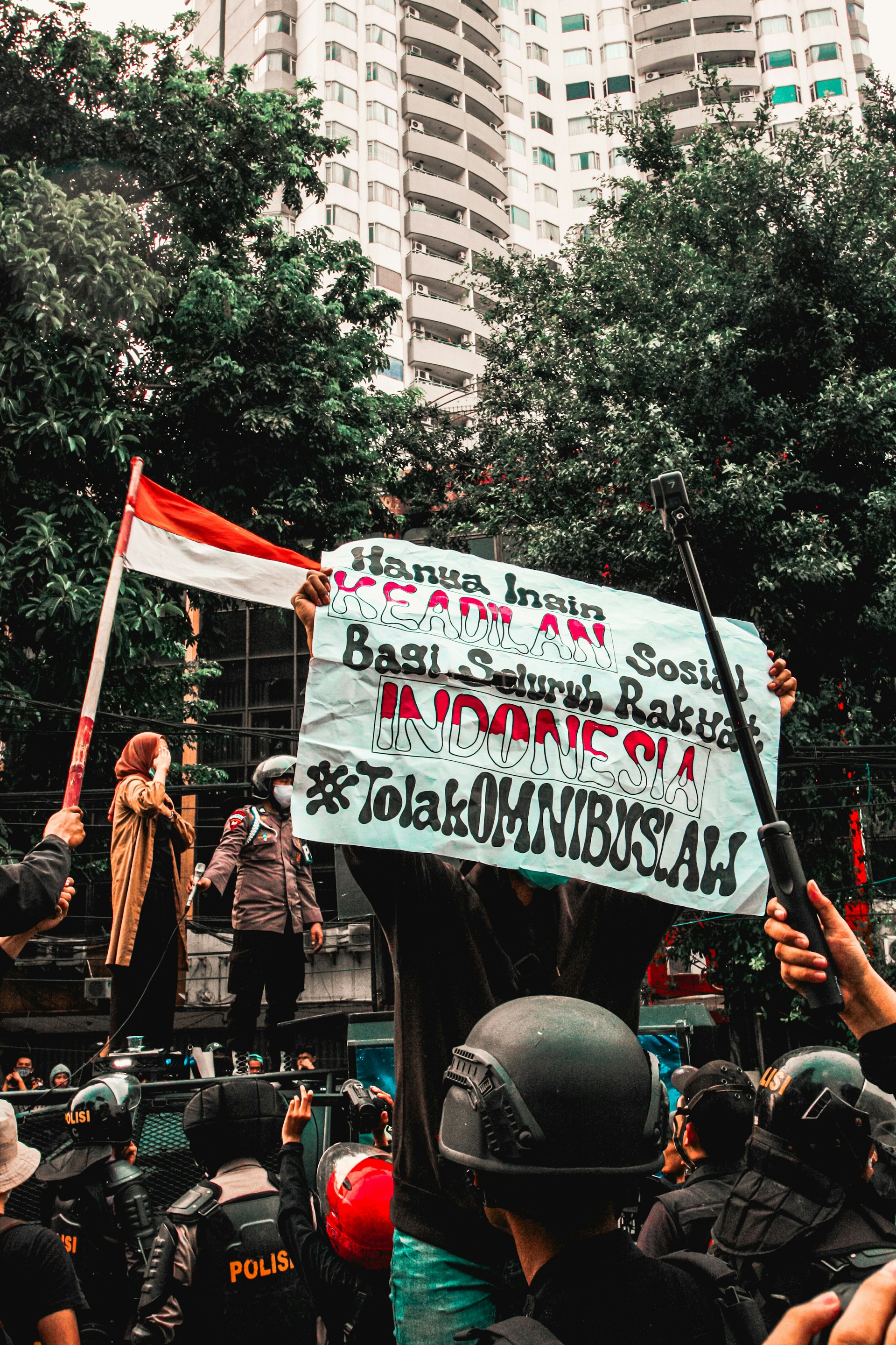 A group of people engaged in a street protest, with one person holding a large banner with text. The crowd includes individuals wearing helmets and uniforms. In the background, there are tall buildings and trees, and a person holds an Indonesian flag.