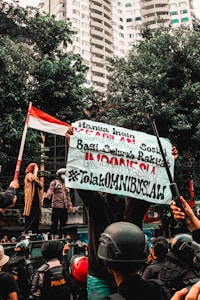 A group of people engaged in a street protest, with one person holding a large banner with text. The crowd includes individuals wearing helmets and uniforms. In the background, there are tall buildings and trees, and a person holds an Indonesian flag.