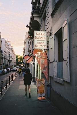 An urban street scene with a supermarket sign reading 'Supermarket Libre Service' above a sidewalk. People are walking along the sidewalk under soft evening light. The street is bordered by tall, narrow buildings, with warm light from setting sun visible in the distance.