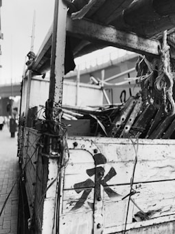 An aged, wooden cart is filled with ropes and tools, showcasing a rustic, industrial setting. The cart displays prominent wear and tear, with peeling paint and visible rust. Overhead, a bridge is seen in the background, while blurred figures walk past on the sidewalk.