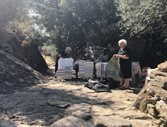 Two women are standing outdoors on a stone pathway surrounded by trees. They are displaying woven textiles, possibly for sale, on a table covered with various colorful cloths in a natural, rustic setting.
