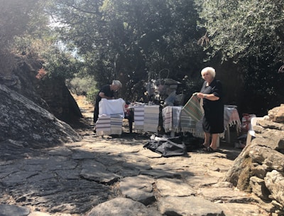 Two women are standing outdoors on a stone pathway surrounded by trees. They are displaying woven textiles, possibly for sale, on a table covered with various colorful cloths in a natural, rustic setting.