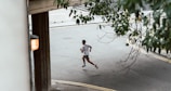 A person is running on a curved road, surrounded by a few bushes and a concrete structure. The street is empty, with a subtle lighting from a lamp attached to the structure.
