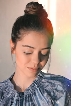 A woman with her hair styled in a bun has her eyes gently closed. She wears a light blue, patterned top, and soft, natural makeup. Sunlight creates a subtle rainbow effect near her face, adding a serene ambiance.