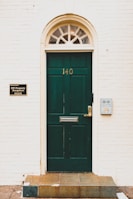 A dark green door with the number 140 is centered within a light-colored brick wall. An arched transom window with multiple panes sits above the door, allowing some light through. To the left of the door, a sign identifies PIP Property Management with contact details, while to the right, there is a modern silver mailbox attached to the wall. The entryway is elevated by a few tiles leading up to the door.