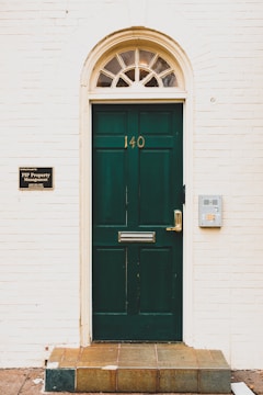 A dark green door with the number 140 is centered within a light-colored brick wall. An arched transom window with multiple panes sits above the door, allowing some light through. To the left of the door, a sign identifies PIP Property Management with contact details, while to the right, there is a modern silver mailbox attached to the wall. The entryway is elevated by a few tiles leading up to the door.