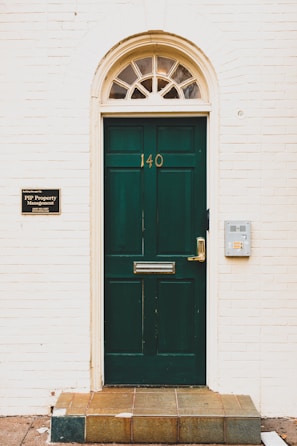 A dark green door with the number 140 is centered within a light-colored brick wall. An arched transom window with multiple panes sits above the door, allowing some light through. To the left of the door, a sign identifies PIP Property Management with contact details, while to the right, there is a modern silver mailbox attached to the wall. The entryway is elevated by a few tiles leading up to the door.