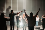 A group of people is gathered in a dimly lit space with raised hands, suggesting an atmosphere of worship or a concert. The lighting creates silhouettes and highlights the participants' outlines, adding to the dramatic effect.