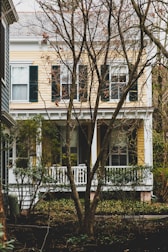 A two-story house with yellow siding and white trim is partially obscured by bare trees in the foreground. The house features several large windows with dark green shutters. There is a small white porch with railings surrounded by shrubs and other greenery.