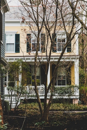 A two-story house with yellow siding and white trim is partially obscured by bare trees in the foreground. The house features several large windows with dark green shutters. There is a small white porch with railings surrounded by shrubs and other greenery.
