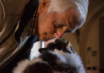 old, bald man in a jacket kissing a cat on the top of its head 