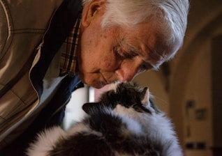 An elderly person gently pressing their forehead against a fluffy cat, conveying a sense of affection and companionship. The setting appears warm and intimate, with soft lighting enhancing the tender moment.