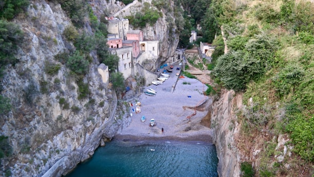 A small beach is nestled between tall rocky cliffs, with a few colorful boats scattered along the pebbled shore. The cliffs are lush with green vegetation, and a series of small, charming buildings with pastel-colored facades are perched against the rock face. The calm, turquoise water gently laps at the shore, creating a serene and secluded atmosphere.