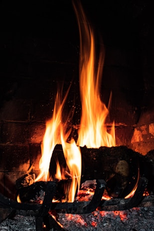 Bright and dynamic flames dance above a set of burning logs in a fireplace, casting warm orange and yellow hues. The wood is cradled in a metal grate, and embers glow beneath, adding a sense of coziness.