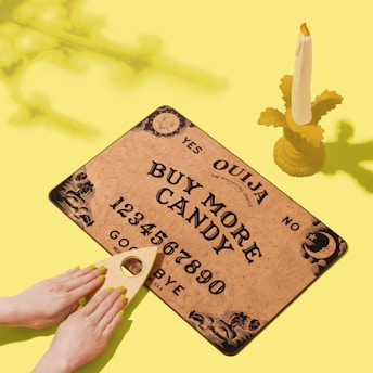 A Ouija board with the message 'Buy More Candy' is placed on a bright yellow surface. A candle with a decorative base stands upright, and two hands with yellow nail polish rest on a planchette. Shadows are cast on the surface, adding depth.