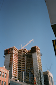 A tall building under construction rises against a clear blue sky. The structure consists of multiple floors with exposed concrete and steel beams. A large crane is positioned at the top, indicating ongoing work. In the foreground, there are other buildings and leafless trees, suggesting a city setting.