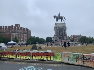 A historical equestrian statue surrounded by colorful graffiti and messages of protest, set in an urban environment with buildings, trees, and people in the background. Tents are pitched on the grass, and there are concrete barriers with slogans and artwork painted on them.