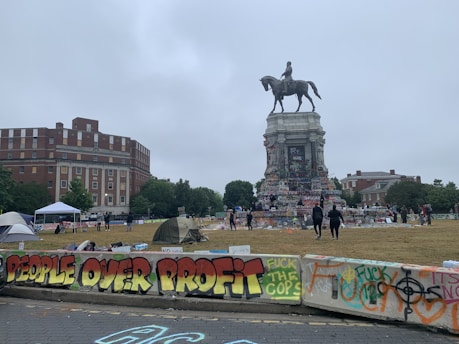 A historical equestrian statue surrounded by colorful graffiti and messages of protest, set in an urban environment with buildings, trees, and people in the background. Tents are pitched on the grass, and there are concrete barriers with slogans and artwork painted on them.