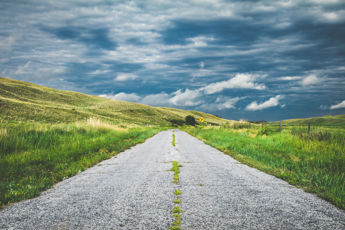 A narrow country road with grass down its middle, between green hills under a cloudy sky