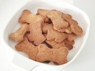 A white bowl filled with numerous bone-shaped brown cookies, arranged in a slightly overlapping manner.