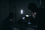 A dimly lit setting with silhouettes of people and filming equipment. One person is operating a camera or recording device with displays emitting soft light. Several stands and a strong vertical light are visible, creating a moody atmosphere.