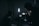 A dimly lit setting with silhouettes of people and filming equipment. One person is operating a camera or recording device with displays emitting soft light. Several stands and a strong vertical light are visible, creating a moody atmosphere.