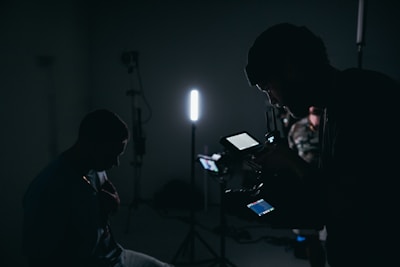 A dimly lit setting with silhouettes of people and filming equipment. One person is operating a camera or recording device with displays emitting soft light. Several stands and a strong vertical light are visible, creating a moody atmosphere.