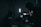 A dimly lit setting with silhouettes of people and filming equipment. One person is operating a camera or recording device with displays emitting soft light. Several stands and a strong vertical light are visible, creating a moody atmosphere.