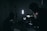 A dimly lit setting with silhouettes of people and filming equipment. One person is operating a camera or recording device with displays emitting soft light. Several stands and a strong vertical light are visible, creating a moody atmosphere.
