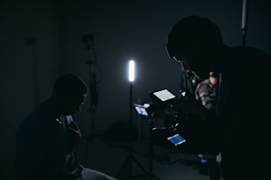 A dimly lit setting with silhouettes of people and filming equipment. One person is operating a camera or recording device with displays emitting soft light. Several stands and a strong vertical light are visible, creating a moody atmosphere.