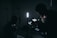 A dimly lit setting with silhouettes of people and filming equipment. One person is operating a camera or recording device with displays emitting soft light. Several stands and a strong vertical light are visible, creating a moody atmosphere.