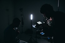 A dimly lit setting with silhouettes of people and filming equipment. One person is operating a camera or recording device with displays emitting soft light. Several stands and a strong vertical light are visible, creating a moody atmosphere.