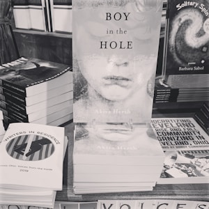 A monochrome image displaying a stack of books on a tabletop with a prominent book titled 'Boy in the Hole' by Akiva Hersh in the center. Other books are surrounding it, including one titled 'Writers in Residence' and another with visible text about community organizing in Cleveland, Ohio.