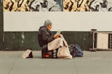 An individual sits on a milk crate on the sidewalk, wrapped in a patterned blanket, and appears to be reading a document. A handwritten cardboard sign is placed on the ground beside them. The background features a mural of abstract black and white figures on a green wall. Nearby bags and a folded board are visible.