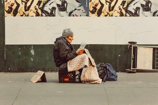 An individual sits on a milk crate on the sidewalk, wrapped in a patterned blanket, and appears to be reading a document. A handwritten cardboard sign is placed on the ground beside them. The background features a mural of abstract black and white figures on a green wall. Nearby bags and a folded board are visible.