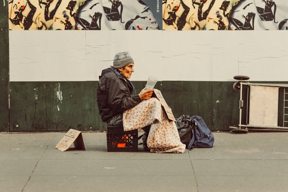 An individual sits on a milk crate on the sidewalk, wrapped in a patterned blanket, and appears to be reading a document. A handwritten cardboard sign is placed on the ground beside them. The background features a mural of abstract black and white figures on a green wall. Nearby bags and a folded board are visible.