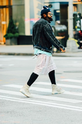 A person wearing a stylish outfit crosses a street. They have a black denim jacket, a dark blue beanie, sunglasses, and beige high-top sneakers. The outfit also includes a patterned headscarf draped around the waist and black leggings underneath.