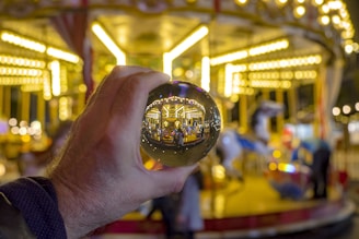 A hand is holding a reflective crystal ball in front of a brightly lit carousel. The background is softly blurred, showcasing bright, warm lights and the carousel's distinct features. Within the crystal ball, the carousel is inverted and more clearly visible, creating an intriguing visual effect.