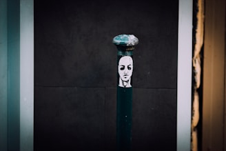 A dark street bollard decorated with a black and white stencil of a serene face in the center. The background consists of dark and light panels, giving a contrast to the artwork on the bollard.