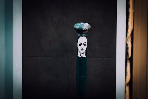 A dark street bollard decorated with a black and white stencil of a serene face in the center. The background consists of dark and light panels, giving a contrast to the artwork on the bollard.