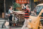 A man stands beside a tricycle loaded with stacks of cardboard, appearing to be a recycling or waste collection setup. Another person in a red and black striped shirt takes a photo using a small camera. There's a bustling street scene with a fast-moving yellow vehicle partially obscuring part of the view. The scene is set in front of a shop displaying decorative signs and plants.
