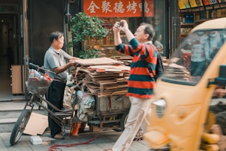 A man stands beside a tricycle loaded with stacks of cardboard, appearing to be a recycling or waste collection setup. Another person in a red and black striped shirt takes a photo using a small camera. There's a bustling street scene with a fast-moving yellow vehicle partially obscuring part of the view. The scene is set in front of a shop displaying decorative signs and plants.