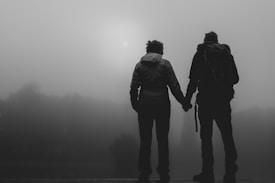 Two silhouetted figures, possibly a couple, stand on a wooden platform holding hands. They gaze towards a foggy, mist-filled landscape with blurred trees and a faint sun visible through the mist.