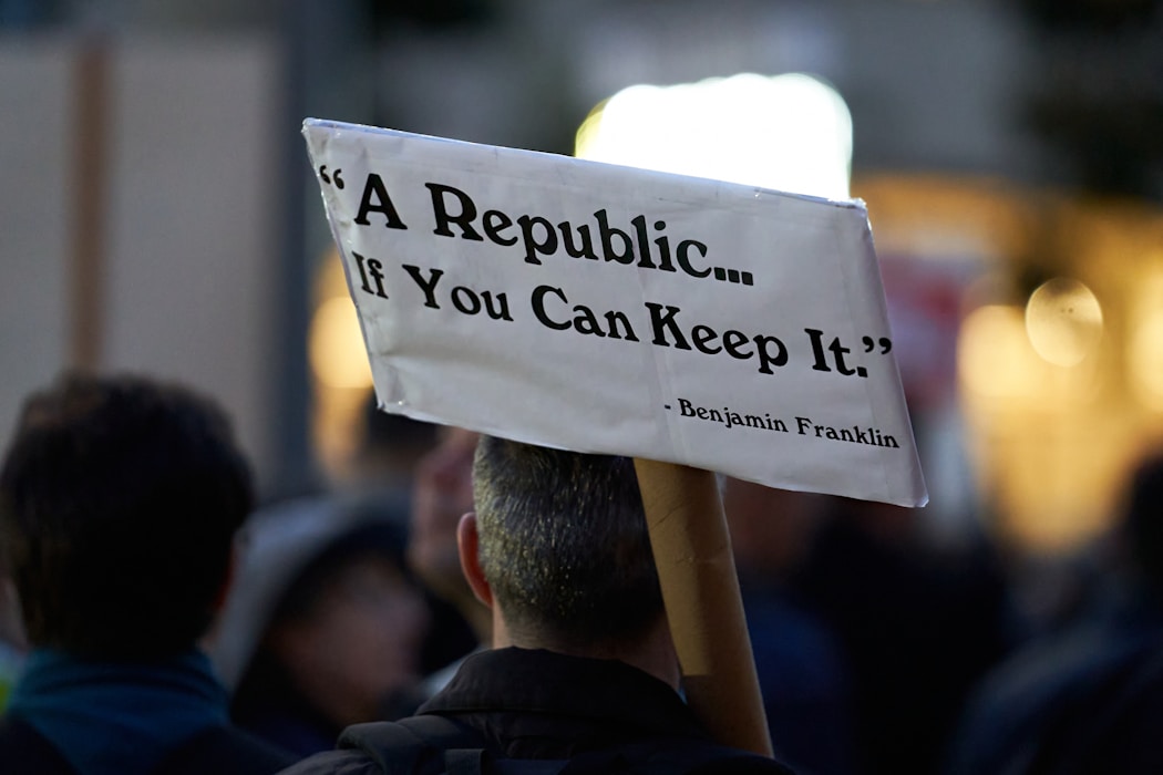 A person is holding up a white sign with the quote 'A Republic... If You Can Keep It.' attributed to Benjamin Franklin. The image is set in a blurred crowd scene, likely at a public gathering or protest.