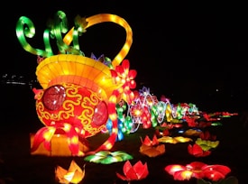 A vibrant, illuminated display featuring a large, colorful structure resembling a decorative, glowing fish with swirling patterns. The fish is surrounded by various brightly lit flowers in multiple colors, set against a dark background, creating a vivid contrast.
