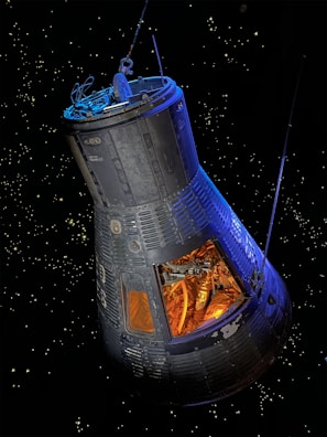 A spacecraft capsule is floating in space against a backdrop of distant stars. The capsule is sleek with visible panels and structure, showing an interior filled with complex machinery and tubing. The light inside the capsule creates a warm, orange glow contrasting with the cold, dark space outside.